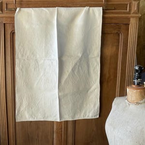 Könnte beinhalten: Ein cremefarbenes, rechteckiges Textil, das an einer Holzstange hängt. Der Stoff hat eine leicht zerknitterte Textur. Der Hintergrund ist ein dunkler Holzschrank. Auf der rechten Seite des Bildes ist eine Schaufensterpuppe zu sehen.