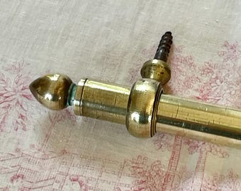 May include: Close-up of a vintage brass curtain rod with a decorative finial. The rod has a cylindrical body with a series of rings and a screw. The finial is a rounded, teardrop shape. The brass has a warm, aged patina.