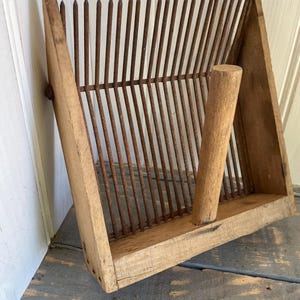 May include: A vintage wooden berry picker with a triangular frame and a series of rusty metal tines. The wooden handle is cylindrical. The frame is made of light-coloured wood, showing signs of age and wear.