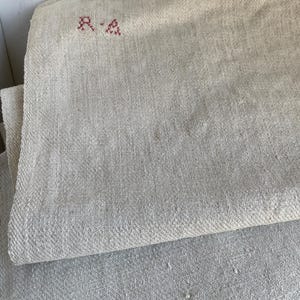 May include: Folded, off-white linen fabric with a woven texture. The top layer has the embroidered letters "R.A" in a reddish-pink color. The fabric appears to be vintage or antique, with a slightly worn appearance. The background is a white, vertical-paneled wall.