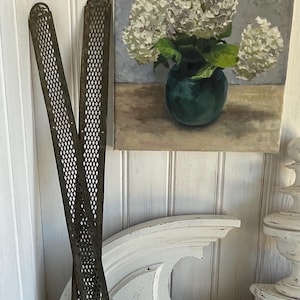 Pode incluir: Duas grelhas decorativas de metal antigas com um padrão de losangos, encostadas a uma parede pintada de branco. Uma pintura de hortênsias brancas num vaso turquesa está acima de um elemento arquitetónico decorativo branco.