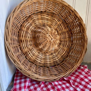 Op de afbeelding: Een grote, ronde, geweven rieten mand met een spiraalvormig ontwerp. De mand is lichtbruin en staat op een rood-wit geruite doek. De mand wordt waarschijnlijk gebruikt om te serveren of op te bergen.