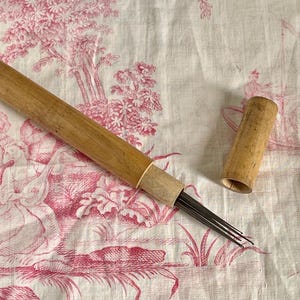 May include: A wooden needle tool with a cylindrical handle and a matching removable cap. The tool features a cluster of thin, sharp metal needles extending from the end of the handle, designed for precision work in various craft applications.