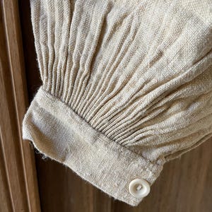 May include: A beige long-sleeve garment cuff featuring vertical pleats gathered into a structured wristband. The cuff is secured with a single, small, round, off-white button. The fabric has a visible, textured, woven appearance.
