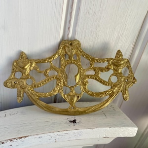 Peut inclure: Poignée de tiroir dorée ornée avec un design de trou de serrure. La quincaillerie décorative présente une base incurvée et des motifs ajourés complexes. La poignée de style vintage est probablement pour une armoire ou une commode.