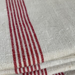 Könnte beinhalten: Nahaufnahme von gefaltetem Leinenstoff in einem natürlichen Off-White-Ton. Auffällige vertikale Streifen in einem satten Rot verlaufen über den Stoff. Die Textur des Leinens ist deutlich sichtbar, was dem Textil einen rustikalen oder Vintage-Look verleiht.