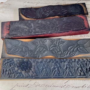 Op de afbeelding: Vier antieke houten drukblokken met gesneden bloemen- en bladerenontwerpen. De blokken zijn rechthoekig met donkergrijze ontwerpen en rode of natuurlijke houten bases. De blokken zijn op elkaar gestapeld.