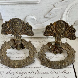 May include: Two matching brass drawer pulls featuring a circular ring handle suspended from a decorative backplate. The backplate displays a central pineapple motif flanked by scrolling foliage, while the ring is adorned with a leaf pattern.