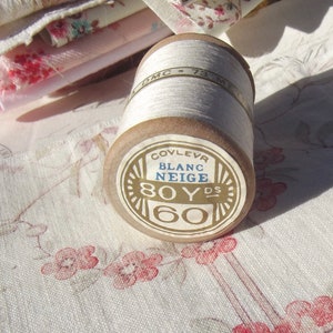 Può includere: Un'bobina di filo bianco con un'etichetta marrone che recita "Couvlevr Blanc Neige 80Yds 60".