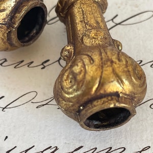 Pode incluir: Close-up de dois objetos decorativos antigos dourados. Um tem formato cilíndrico com base alargada e detalhes ornamentados. O outro tem formato arredondado. Ambos têm interior oco. Os objetos estão sobre uma superfície de papel.