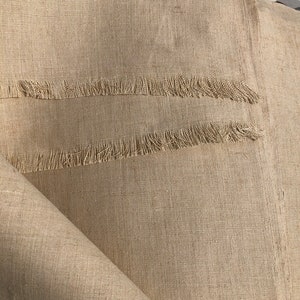 Op de afbeelding: Close-up van beige linnen stof met een gefranjerde rand. De stof is gevouwen en onthult de textuur en het weefsel van het materiaal. De neutrale kleur en het eenvoudige ontwerp suggereren een rustieke of minimalistische esthetiek.