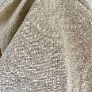 Könnte beinhalten: Nahaufnahme eines beigefarbenen Leinenstoffs mit strukturiertem Gewebe. Der Stoff ist gefaltet und zeigt die natürlichen Farbvariationen und die detaillierte Textur des Materials. Das Bild hebt die organische Optik des Leinens hervor.