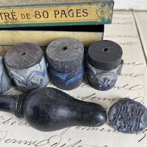 Pode incluir: Uma variedade de ferramentas de escrita antigas, incluindo carimbos de tinta e uma caneta, em tons de preto, azul e cinza. Uma caixa vintage com o texto "TRE DE 80 PAGES" está ao fundo. Os itens estão dispostos em um papel com escrita.