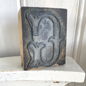 May include: A vintage wooden letterpress block featuring the letter "D" in a decorative, ornate font. The block is dark gray with a weathered appearance, mounted on a wooden base. The block is displayed on a white shelf.