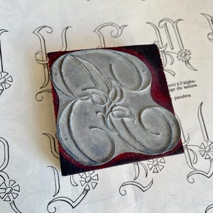 Może przedstawiać: Zabytkowy blok drukarski z literą 'B' w stylu vintage. Metalowa litera jest zamontowana na ciemnoczerwonym i czarnym drewnianym bloku. Tło przedstawia kartkę papieru z podobnymi wzorami liter.