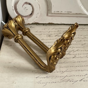 Może przedstawiać: Para ozdobnych, złotych uchwytów do zasłon. Uchwyty mają szczegółowy, liściasty wzór i są zamontowane na śrubie. Tło obejmuje kawałek antycznego papieru z odręcznym pismem i biały, dekoracyjny element architektoniczny.