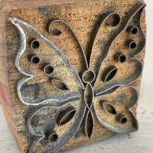 Könnte beinhalten: Ein Vintage-Schmetterlingsstempel aus Holz und Metall. Das Schmetterlingsdesign besteht aus Metall und ist auf einem rechteckigen Holzblock montiert. Das Holz hat ein natürliches, gealtertes Aussehen mit sichtbarer Maserung und Textur. Der Metallschmetterling hat eine detaillierte Kontur.