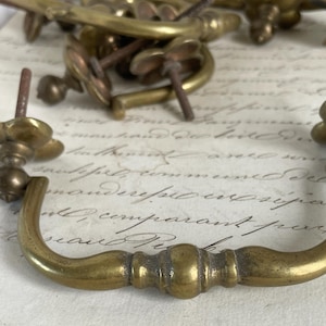 Op de afbeelding: Close-up van antieke messing lade grepen en knoppen. De hardware heeft een vintage esthetiek, met een gebogen handvat en decoratieve bloemaccenten. Het messing heeft een warme, verouderde patina.