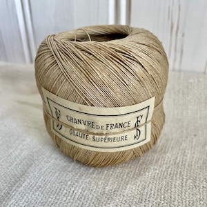 May include: A ball of natural-colored twine with a label wrapped around it. The label reads "Chanvre de France Qualite Superieure" in black text. The twine is tightly wound and appears to be made of natural fibers.