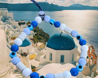 May include: A beaded bracelet featuring a mix of blue, white, and gray beads. The bracelet includes round beads, cylindrical beads, and beads with an eye design. A blue cord is used to string the beads together.
