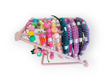 May include: A collection of colorful bracelets displayed on a pink metal rack. The bracelets feature various designs, including beads, braided cords, and decorative charms. Colors include pink, green, orange, and blue. The bracelets are suitable for children.