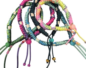 May include: A collection of colorful paracord wrist lanyards. Each lanyard features a braided design in various colors, including pink, green, blue, and black, with wooden beads and adjustable cords. Some lanyards have letter beads.