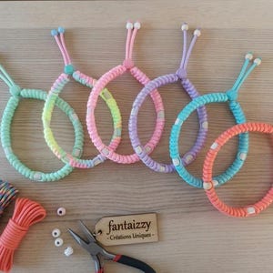 Könnte beinhalten: Eine Sammlung farbenfroher, handgefertigter Armbänder in Rosa, Blau, Grün und Orange. Die Armbänder sind aus geflochtenem Kordel gefertigt und haben einen Schiebeknotenverschluss. Das Bild zeigt auch Kordelspulen, Perlen und eine Zange.