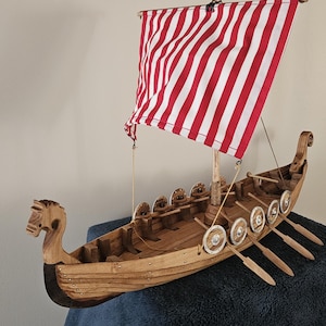Könnte beinhalten: Ein hölzernes Wikingerschiff-Modell mit einem geschnitzten Drachenkopf am Bug, einem Mast mit einem rot-weiß gestreiften Stoffsegel und mehreren hölzernen Rudern an den Seiten. Der Rumpf ist aus hellbraunem Holz mit sichtbarer Maserung gefertigt.