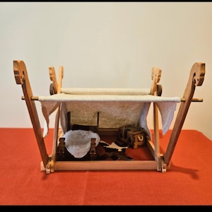 Könnte beinhalten: Ein Miniatur-Wikinger-Schiffbett aus Holz mit einem Segeltuchdach. Der Bettrahmen besteht aus hellem Holz mit dekorativen Schnitzereien an den senkrechten Stützen. Im Inneren des Bettes befinden sich Miniaturgegenstände, darunter ein Fell und eine kleine Truhe.