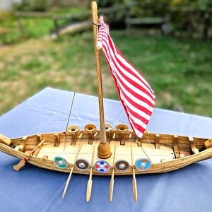 Op de afbeelding: Een houten model van een Vikingschip met een lichtbruine romp, een uitgesneden drakenkop bij de boeg en een gekrulde staart bij de achtersteven. Het schip heeft een centrale mast met een rood-wit gestreept zeil en kleine ronde schilden.