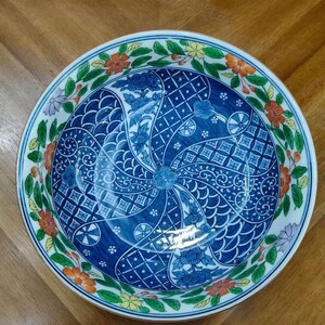 May include: A round ceramic bowl with a detailed blue and white floral design. The bowl's interior features intricate patterns, while the rim is adorned with colorful flowers and green leaves. The bowl is set against a wooden surface.