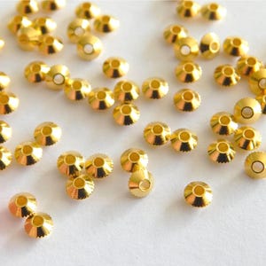 Könnte beinhalten: Eine Ansammlung kleiner, goldfarbener Metallperlen. Jede Perle hat eine abgerundete, kegelförmige Form mit einem Loch oben und einem kleineren Loch unten. Die Perlen sind auf einer weißen Oberfläche verstreut.