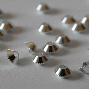 Könnte beinhalten: Eine Ansammlung kleiner, silberfarbener, kegelförmiger Perlen, die auf einer hellen Oberfläche verstreut sind. Jede Perle hat einen ausgestellten, geriffelten Rand und ein kleines Loch oben. Die Perlen sind reflektierend.
