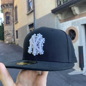Könnte beinhalten: Schwarze Baseballkappe mit dem Logo der New York Yankees in Weiß, verziert mit kleinen, perlenartigen Akzenten. Die Kappe hat einen flachen Schirm und eine strukturierte Krone, geeignet für den Freizeitgebrauch.