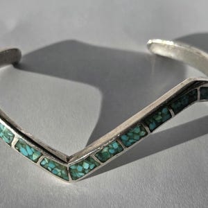 May include: A silver-toned cuff bracelet with a V-shaped design. The bracelet is inlaid with small, rectangular turquoise stones. The open-ended design allows for easy wear. The bracelet is displayed on a light gray surface.