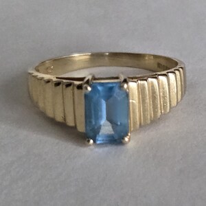 Könnte beinhalten: Ein goldener Ring mit einem rechteckigen, hellblauen Edelstein. Das Band des Rings hat ein geripptes Design. Der Edelstein ist in einem klassischen, minimalistischen Stil gefasst. Der Ring ist wahrscheinlich aus Gold.