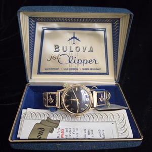 May include: Vintage Bulova Jet Clipper watch in its original blue presentation box. The watch has a gold-tone case and a dark face with gold hands and numerals. The watch band features blue accents with Masonic symbols. The box has the Bulova logo and text.
