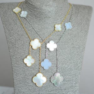 May include: Three necklaces with clover-shaped pendants on a gray mannequin. The necklaces have gold and silver chains. The pendants are white, light blue, and iridescent, with gold or silver borders. The necklaces are a jewelry item.