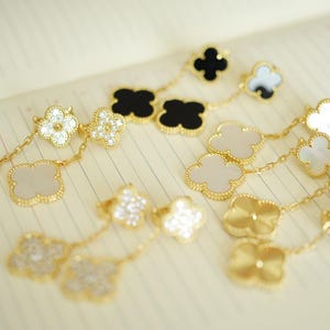 May include: A collection of gold-toned drop earrings featuring four-leaf clover motifs. The earrings include various designs with black, white, and iridescent inlay, as well as pavé-set clear stones, all connected by delicate gold-toned link chains.