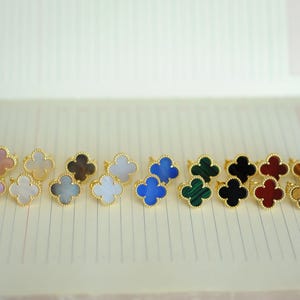 May include: A collection of gold-toned rings, each featuring a four-leaf clover design in various colors. Colors include pink, white, brown, blue, green, black, red, and tiger's eye. The rings are displayed on a lined surface.