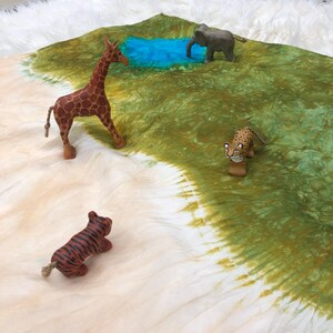 May include: A wooden giraffe, tiger, leopard, and elephant figurine set on a green and white tie-dye fabric background. The figurines are arranged around a blue pool of water.