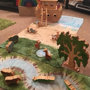 May include: A wooden toy ark with a ladder and two figures standing on the roof. The ark is surrounded by a green and blue play mat with wooden animal figurines, including giraffes, elephants, zebras, and crocodiles.