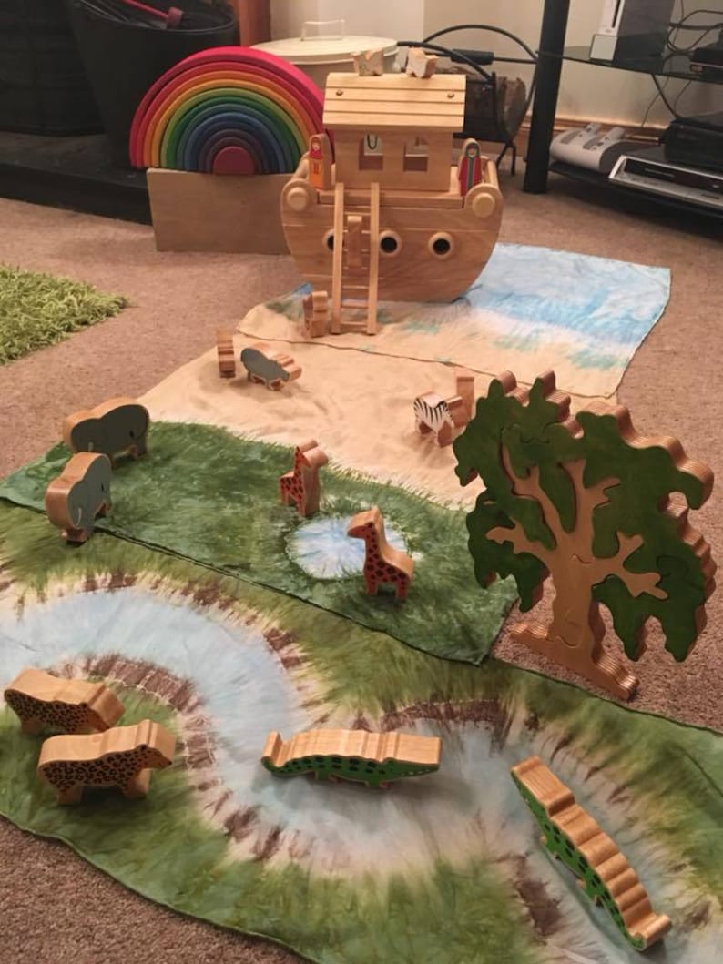 May include: A wooden toy ark with a ladder and two figures standing on the roof. The ark is surrounded by a green and blue play mat with wooden animal figurines, including giraffes, elephants, zebras, and crocodiles.