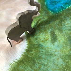 May include: A wooden elephant figurine with a raised trunk, standing on a white and green tie-dye fabric background.