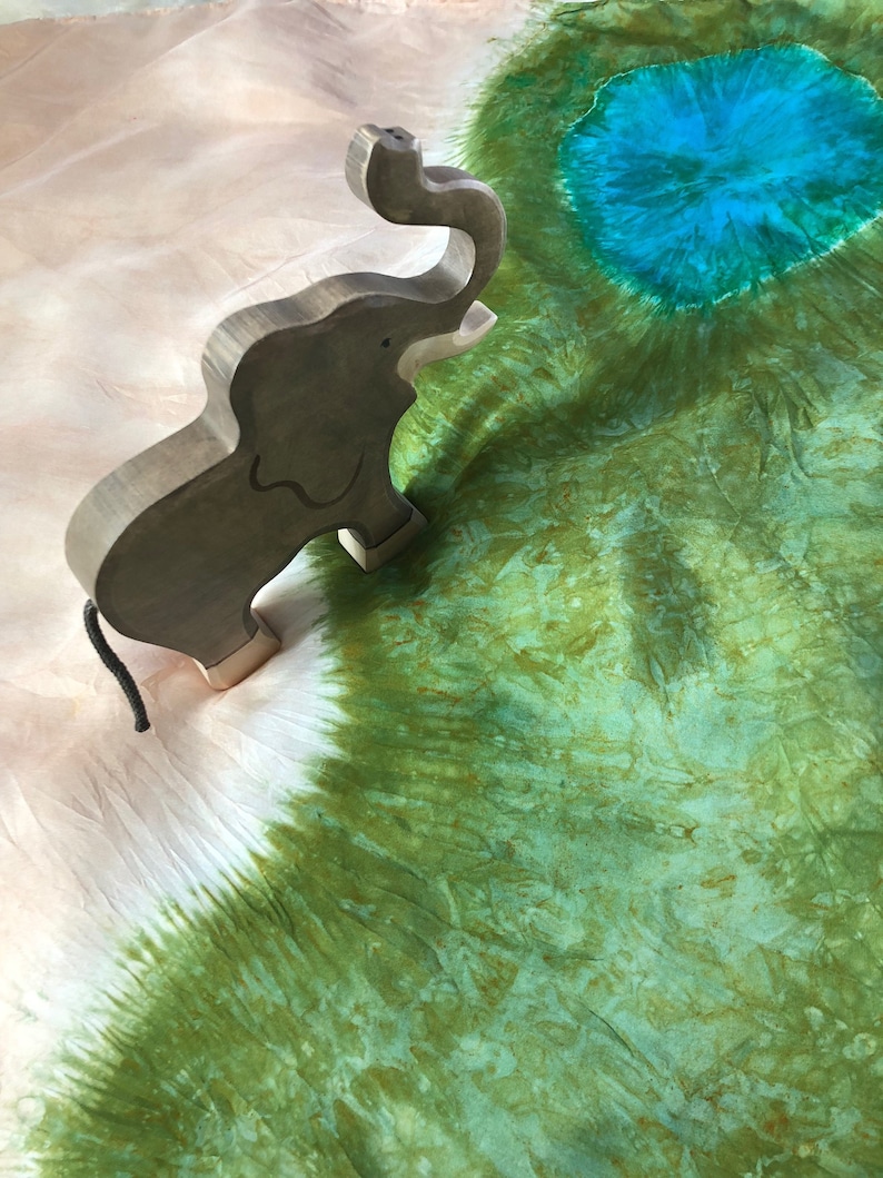 May include: A wooden elephant figurine with a raised trunk, standing on a white and green tie-dye fabric background.