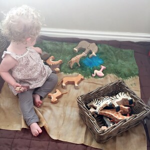May include: A child plays with wooden animal figurines on a green and brown play mat. The figurines include a lion, giraffe, elephant, zebra, and flamingo. The child is holding a wooden block in their hand.