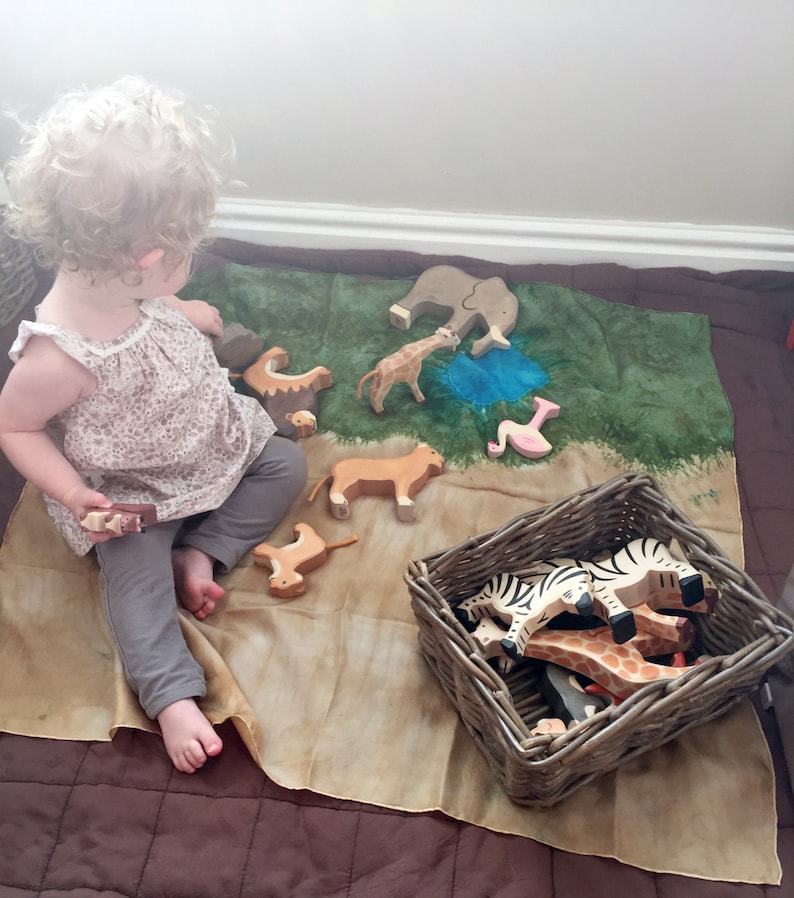 May include: A child plays with wooden animal figurines on a green and brown play mat. The figurines include a lion, giraffe, elephant, zebra, and flamingo. The child is holding a wooden block in their hand.