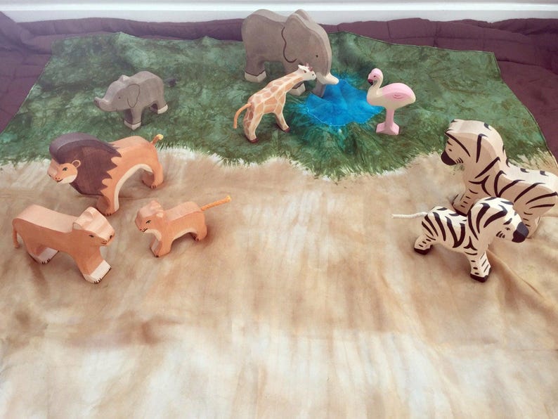 May include: A set of wooden animal figurines, including a lion, a giraffe, a zebra, an elephant, a flamingo, and two lion cubs. The figurines are arranged on a green and brown fabric background.