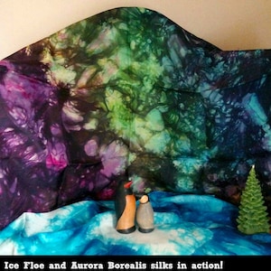 May include: Two wooden penguin figurines stand on a blue fabric background. The fabric is patterned with dark blue, green, and purple swirls. The penguins are facing each other. The text "Ice Floe and Aurora Borealis silks in action!" is at the bottom of the image.