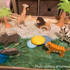 May include: A wooden play set with a variety of animal figurines, including a giraffe, zebra, tiger, elephant, and lion. The set also includes two palm trees and a green and brown play mat.